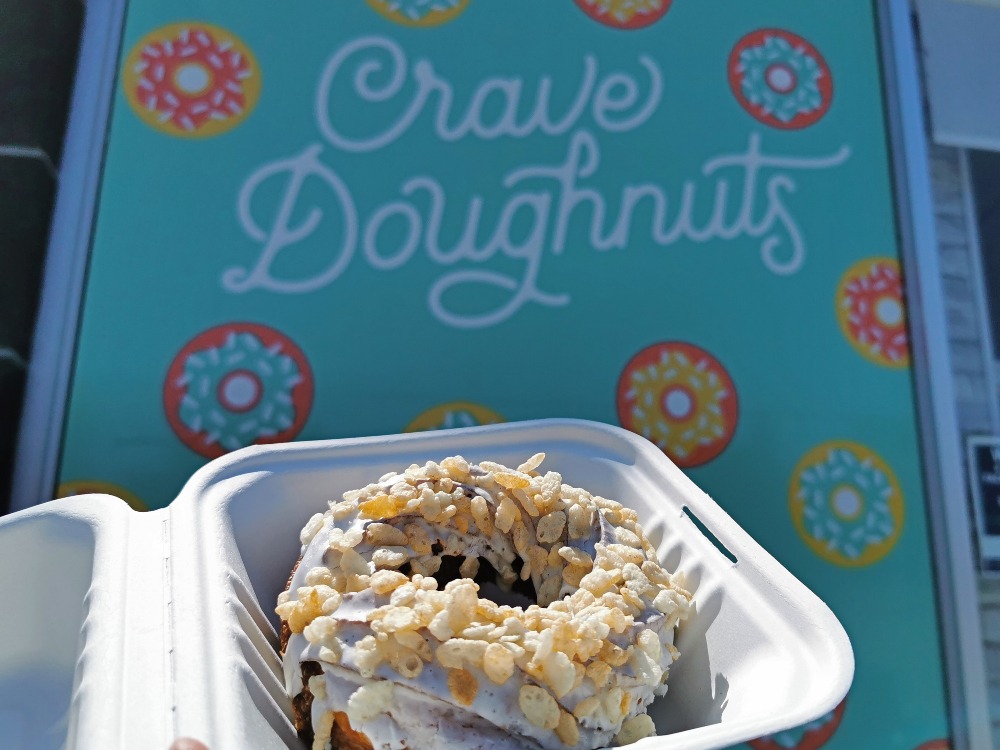 Crave Doughnuts Rice Krispy Cruller in a small container, held up in front of the store's sign