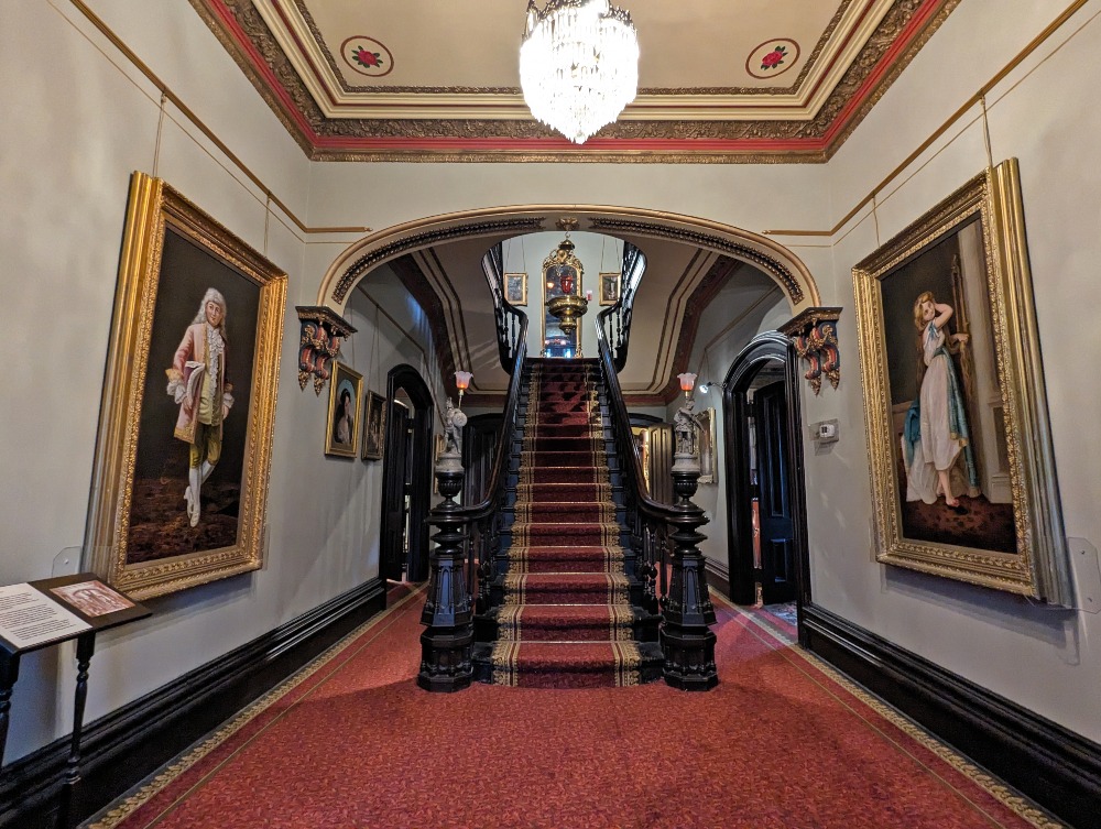 Entryway of Glanmore National Historic Site with grand wooden staircase at the centre, painted ceiling and two large paintings on the walls.