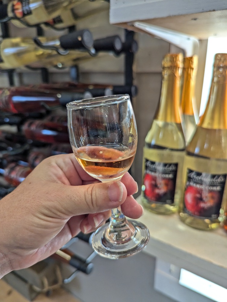 Hand holding up a small glass of wine at Archibald Orchards & Estate Winery, with large bottles of wine in the backround.