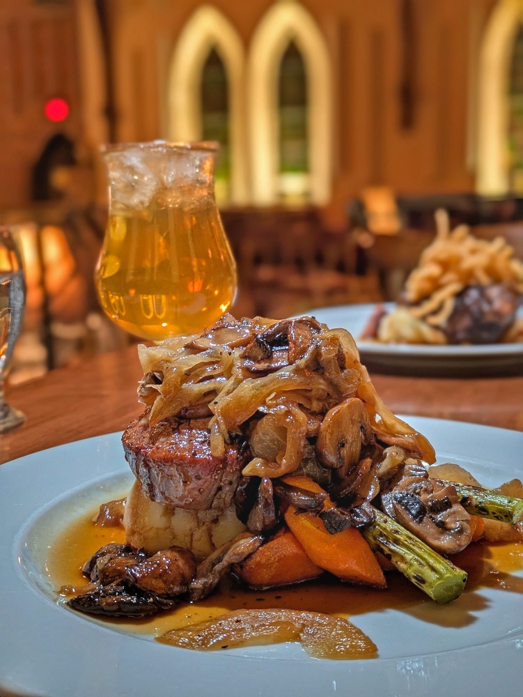 Plate of steak topped with onions and sitting on top of vegetables. In the background are old church windows.