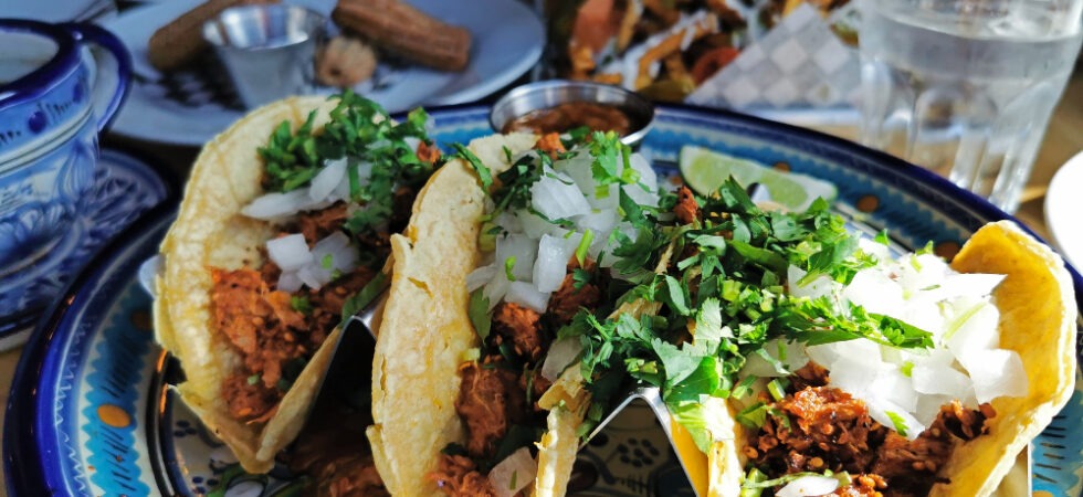 Plate of three tacos in the forefront with other Mexican dishes in the background.