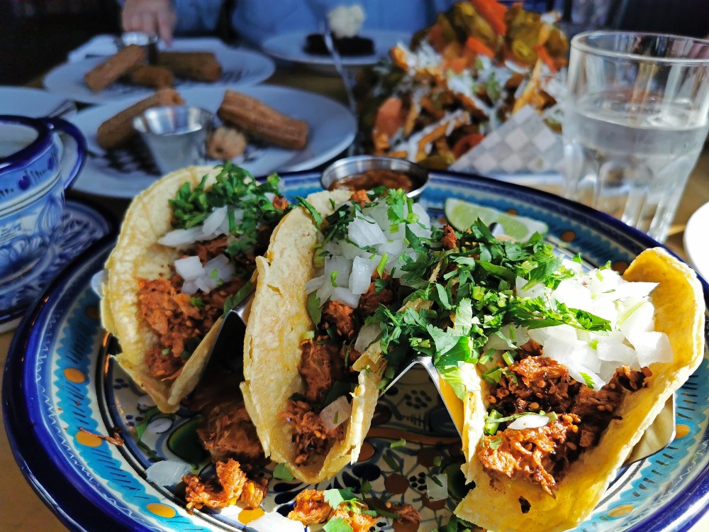 Plate of three tacos in the forefront with other Mexican dishes in the background.