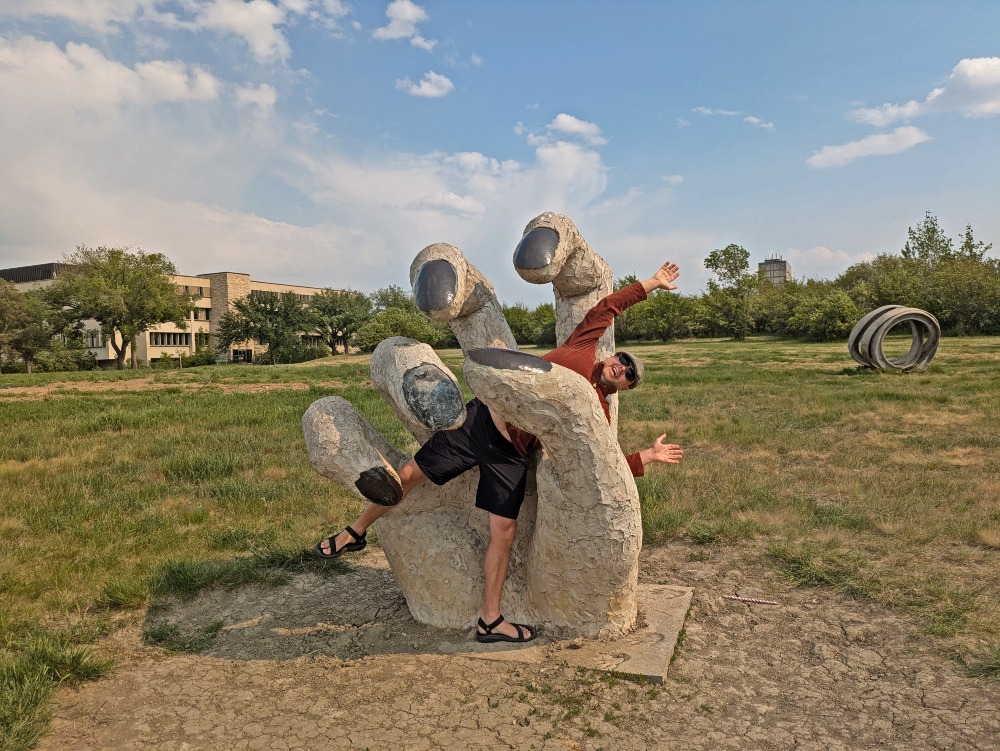 Man poising with large hand sculpture like the hand is holding him, at the Sculpture Park in Saskatoon