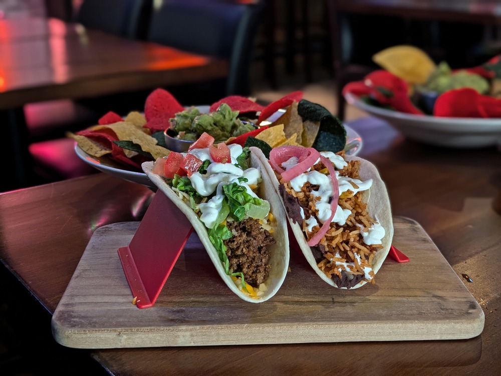 Two tacos with a plate of guacamole and chips in the background