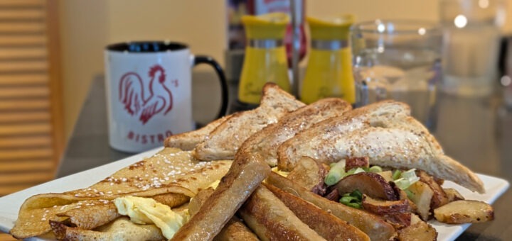 Big breakfast plate with sausages, Finnish pancakes, eggs and more at the Rooster's Bistro, with coffee cup with a red rooster on it in the background