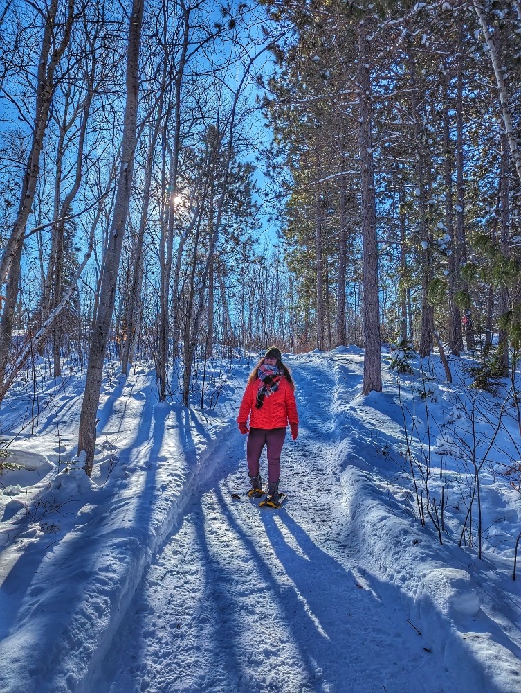 Me snowshoeing in Kivi Park in Sudbury in the winter. The trail is lined with towering pines with blue sky peeking through.