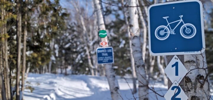 fat biking trail in Kivi Park with sign on a tree in the foreground. Snow covering the ground of the trail and through the forest that lines the trail.