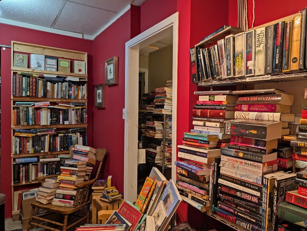 Red room full of books on shelves and piled on chairs at By The Book PTBO