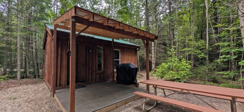 A cabin at Brennan's Hill campground that's surrounded by forest, has a picnic table and bbq as well as a covered deck