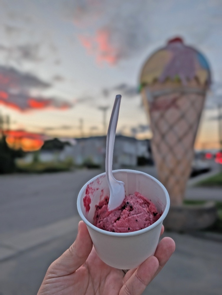 Hand holding up a cup of ice cream in from of a massive ice cream cone at sunset